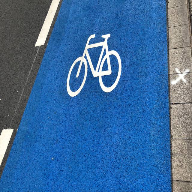 Ein blauer Schutzstreifen auf einer Straße mit Fahrrad (in weiß)