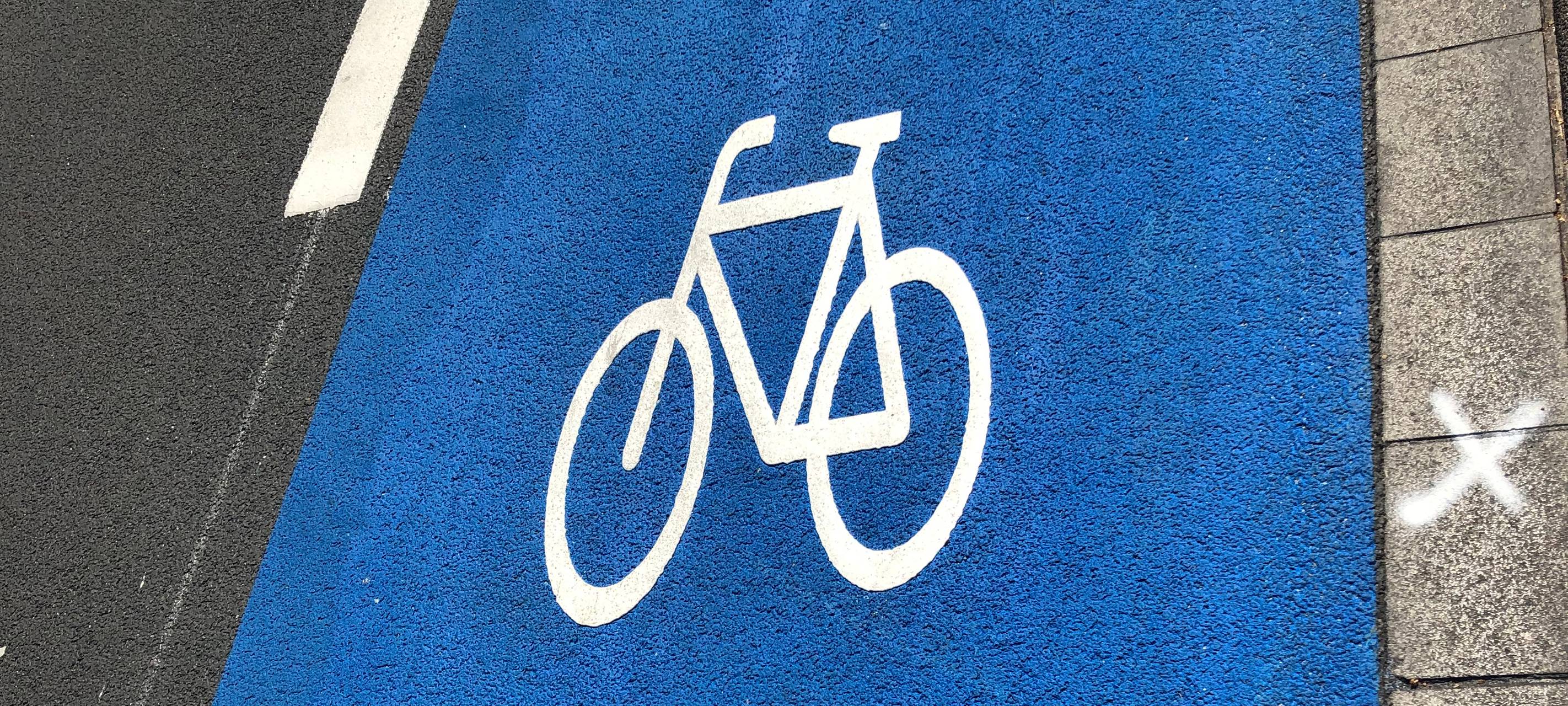 Ein blauer Schutzstreifen auf einer Straße mit Fahrrad (in weiß)