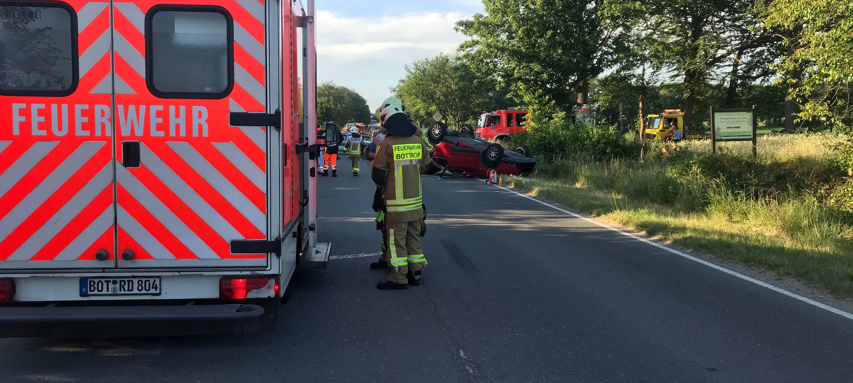 Ein Rettungswagen steht im Vordergrund, im Hintergrund sieht man ein rotes Auto auf dem Dach liegen, davor Feuerwehrleute.