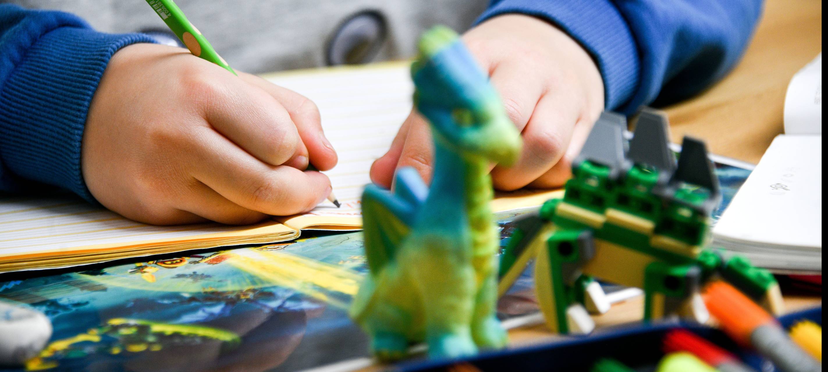 Ein Kind schreibt in ein Heft, vor ihm liegen Spielsachen