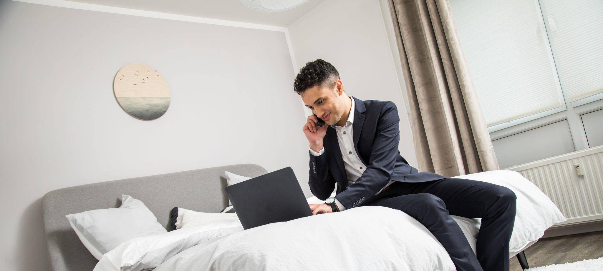 Ein Mann im Anzug sitzt auf einem Bett vor seinem Laptop